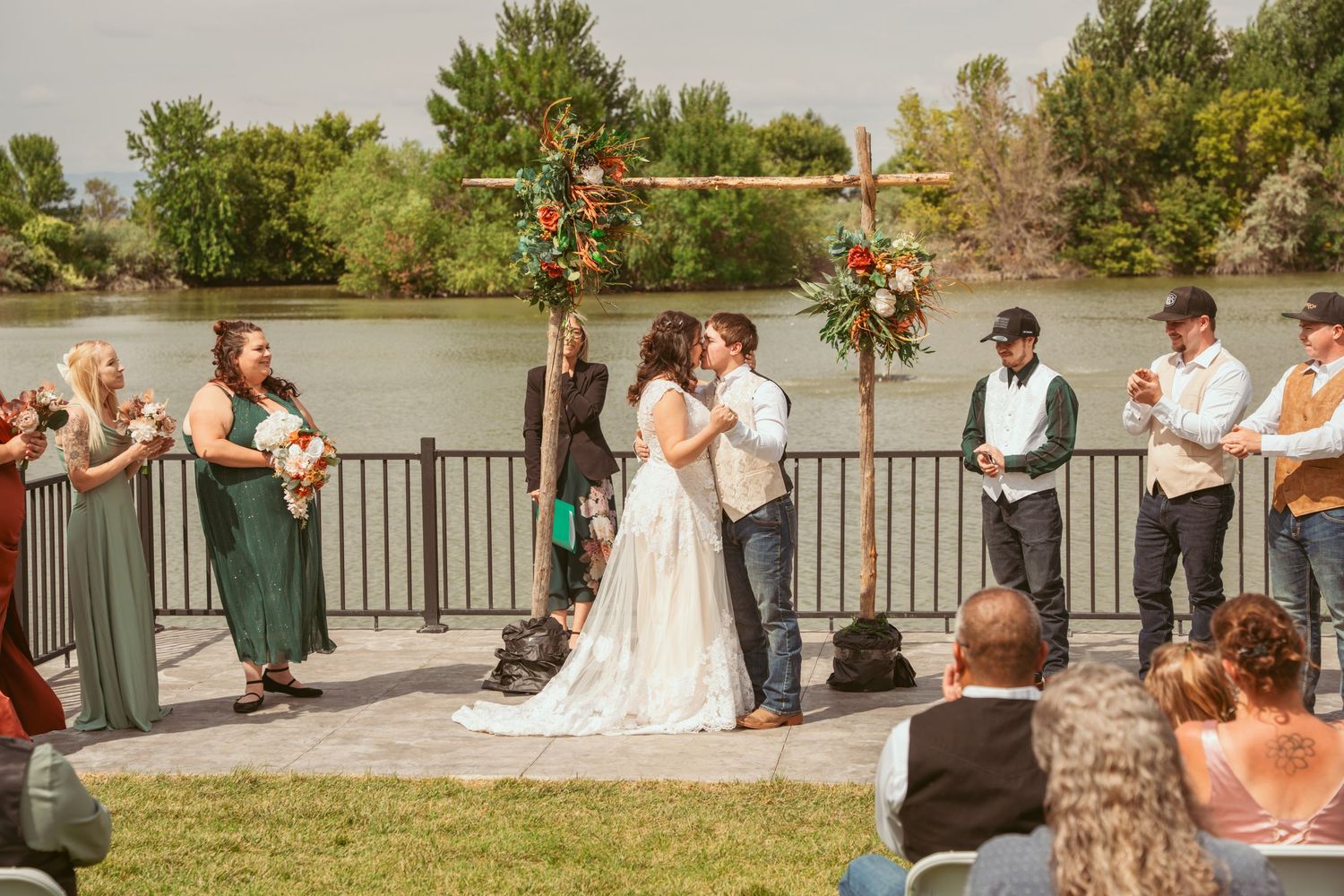 A couple stands in a sunlit meadow wearing wedding attire surrounded by evergreen trees during golden hour.