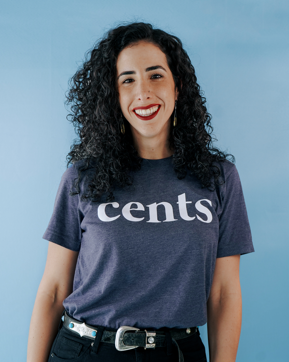 A headshot of a person with curly hair wearing a navy t-shirt with 'cents' text poses against a blue background.