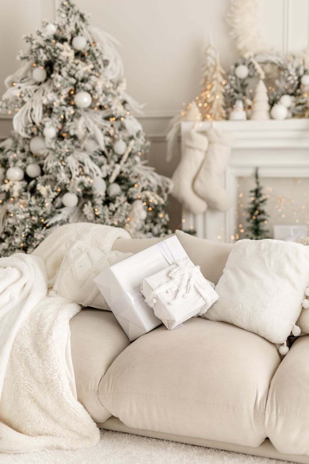 Cozy white Christmas living room with flocked tree, stockings on mantel, wrapped gifts and plush pillows on sofa.