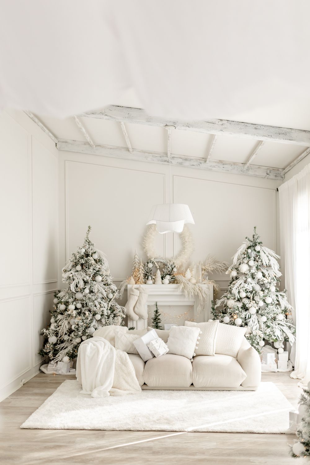 Elegant white Christmas decor featuring two flocked trees, plush sofa, and fireplace mantel in monochromatic winter theme.