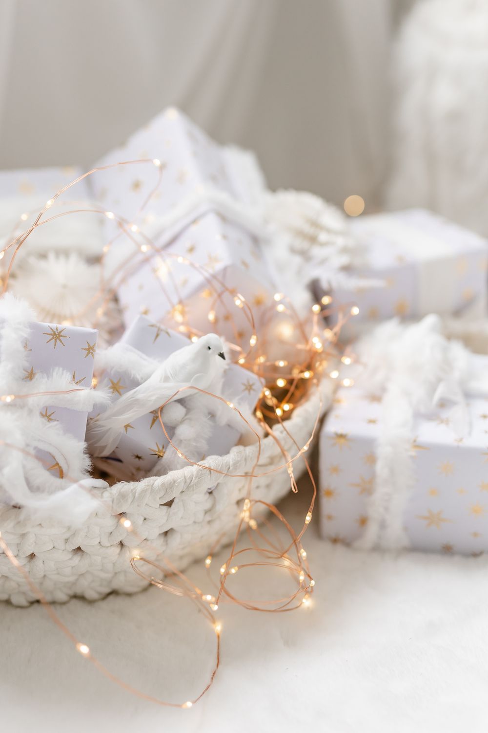 White wrapped holiday gifts with gold star pattern arranged in woven basket with delicate twinkle lights creating cozy ambiance.