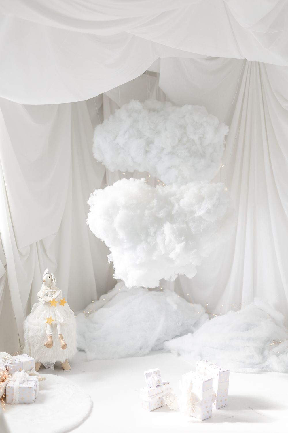 White fluffy cotton cloud decorations hanging above a dreamy scene with lanterns and wrapped gifts in monochromatic decor.