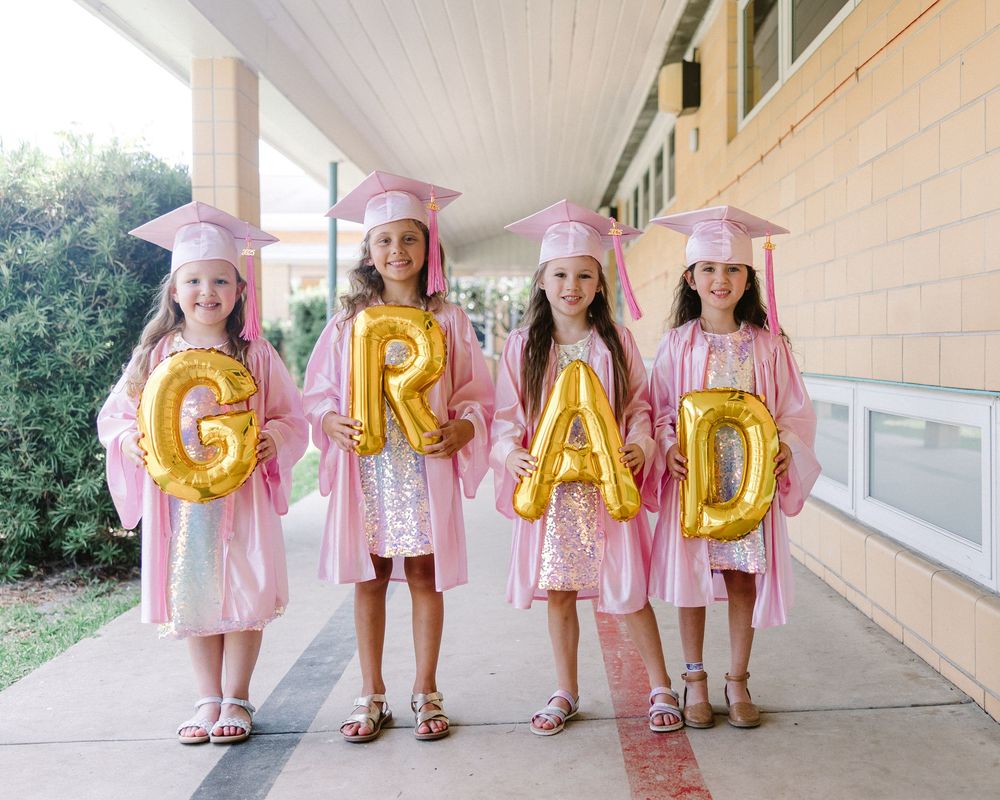 Young graduates in pink caps and gowns hold gold letter balloons spelling out GRAD while standing in a covered walkway.