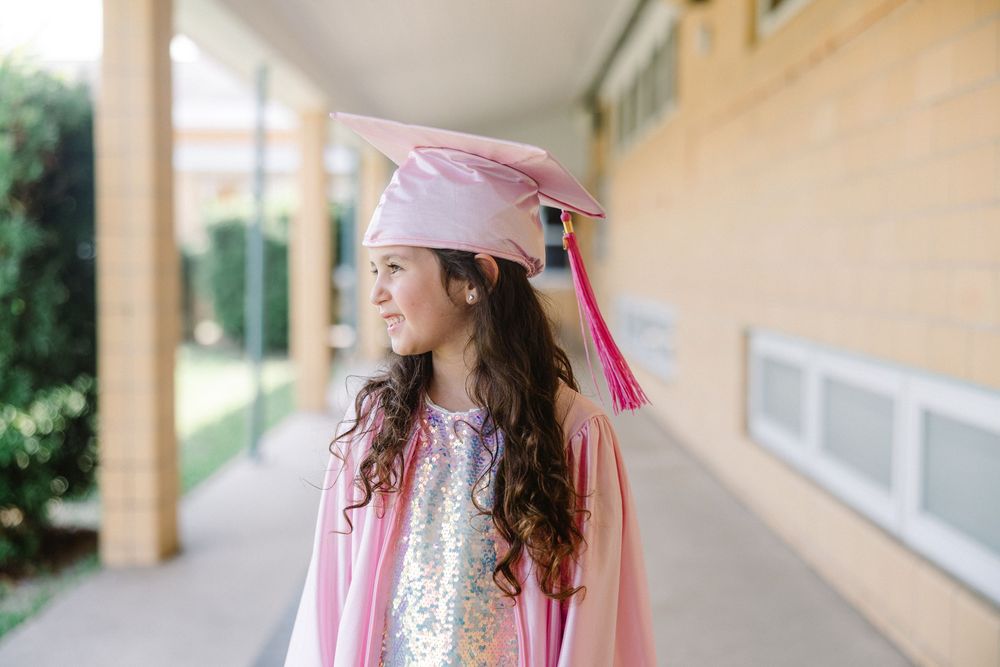 A young graduate in white cap and pink dress poses confidently in multiple frames along a sunlit corridor.