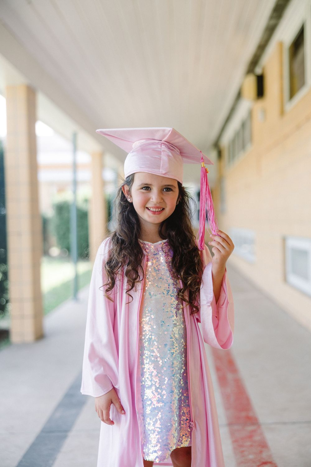 Young girl wearing a pink jacket and sequined top smiles while walking along a covered outdoor walkway at school.