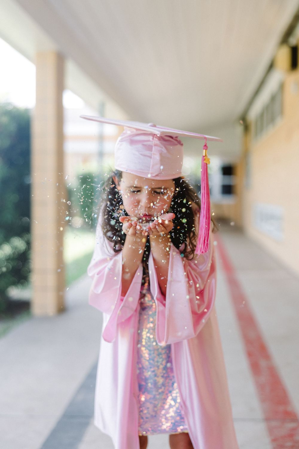 Graduate in pink cap and gown blows glitter while celebrating in a covered walkway.