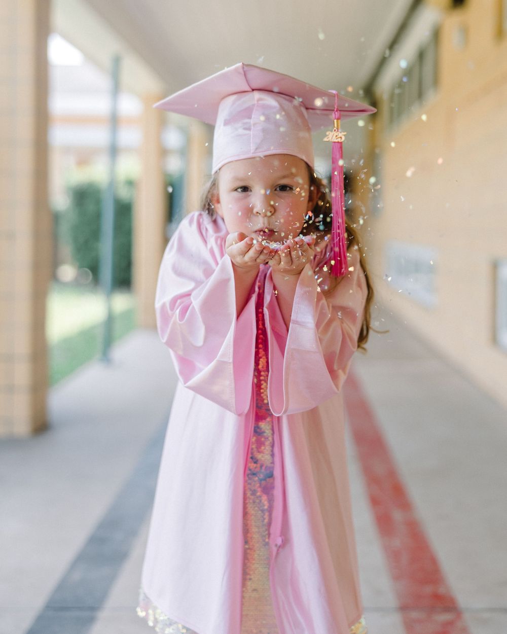 Three views of a graduate in pink cap and gown walking through a sunlit hallway corridor.