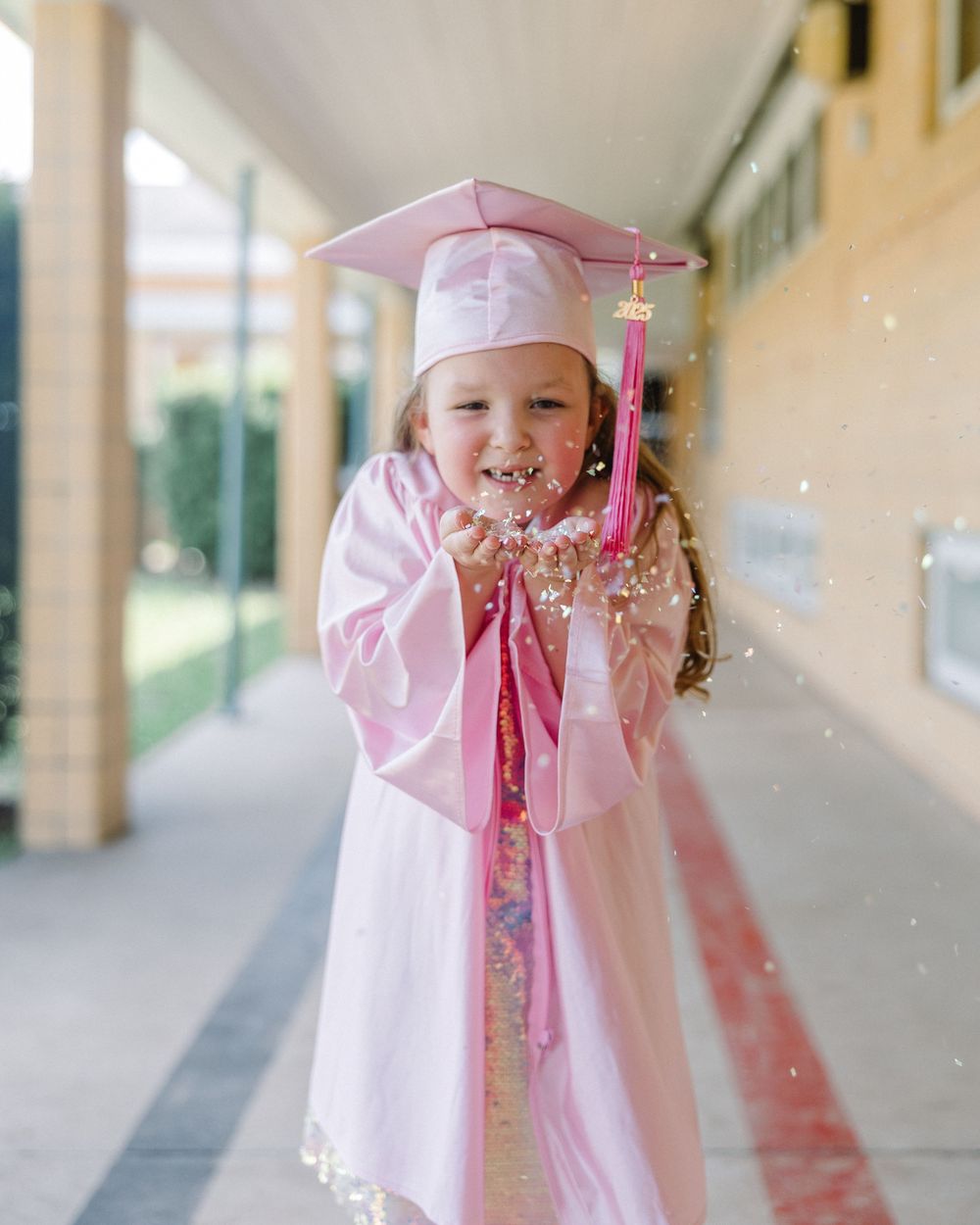 Pink-clad graduate in cap and tassel blows glittery confetti in school hallway during celebratory moment.