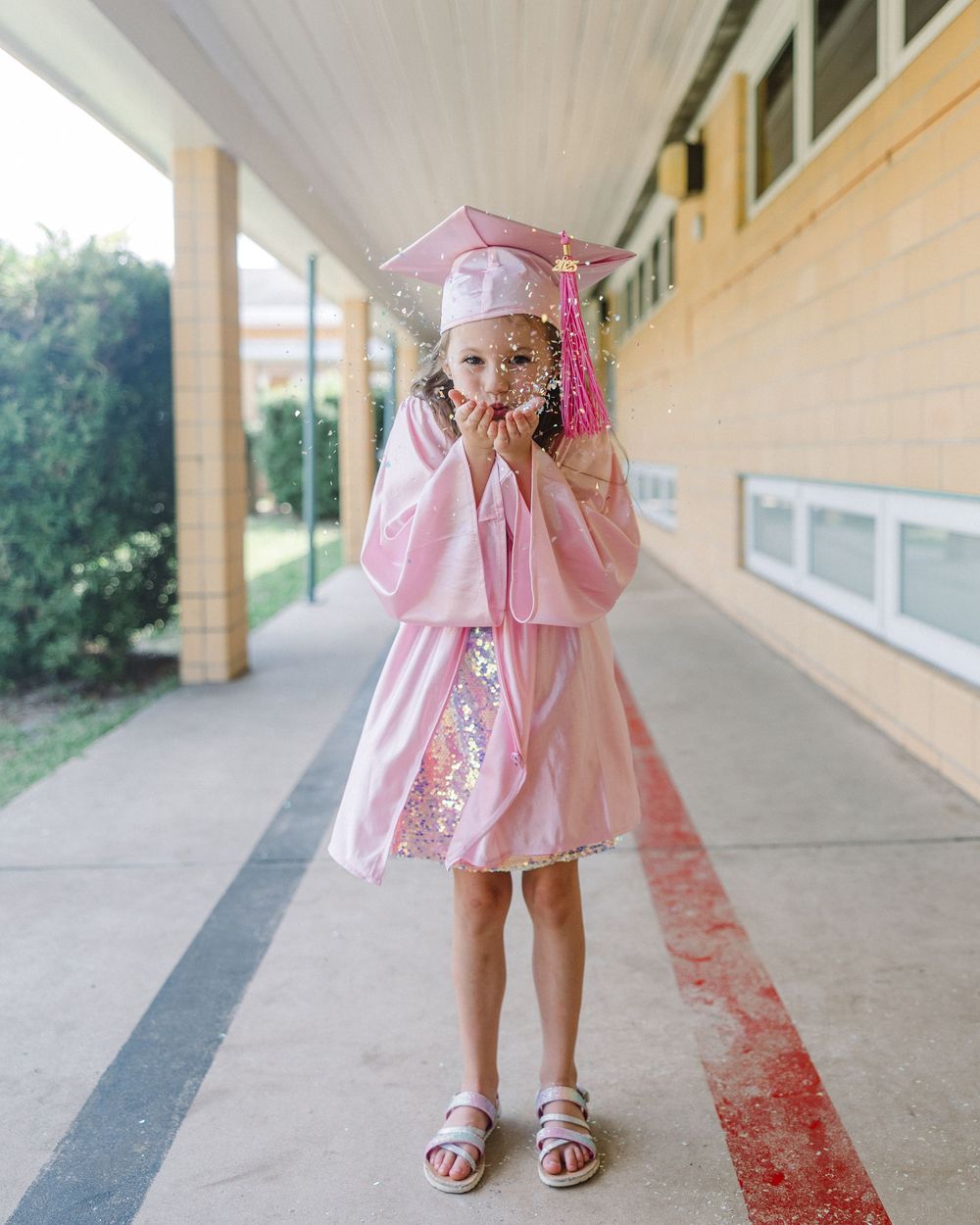 A person wearing a pink graduation gown with matching cap stands in a covered walkway outside a building.