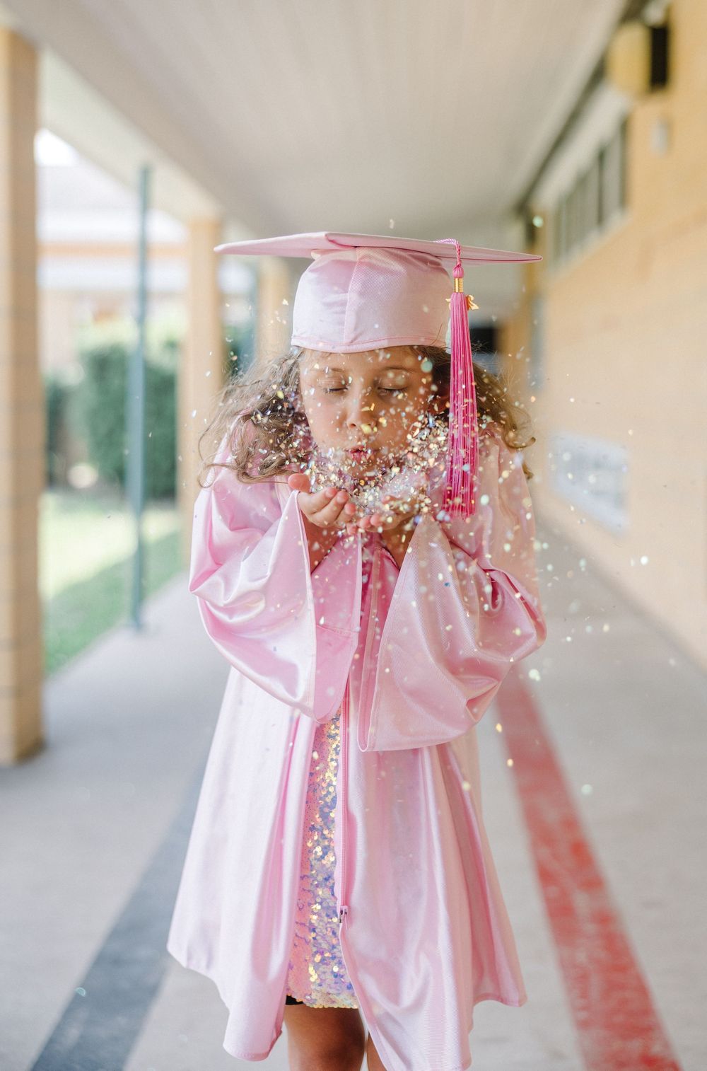 A graduate in pink cap and gown blows glitter while standing in a school hallway during celebration photos.