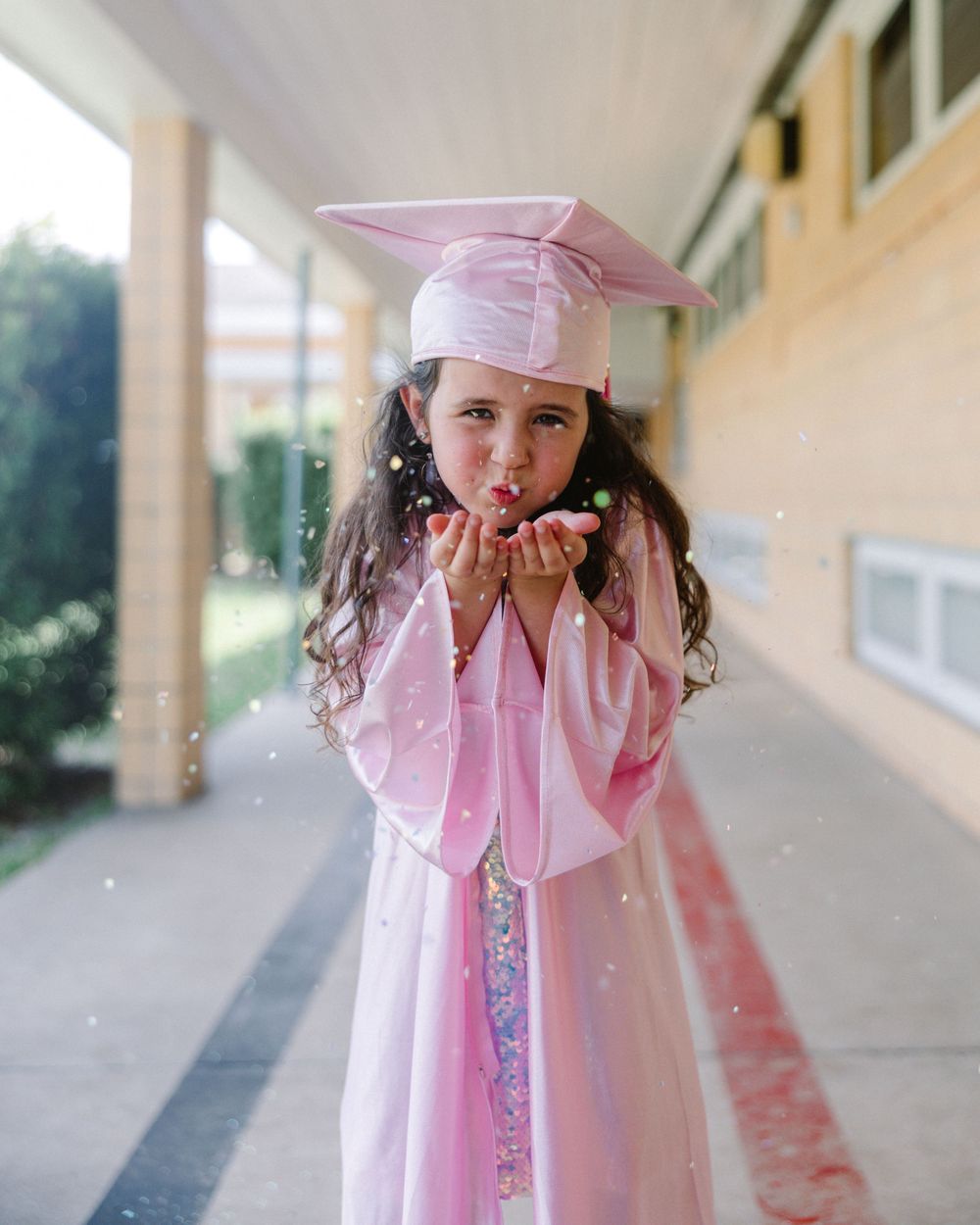 Graduate wearing pink cap and gown blows sparkly confetti in covered hallway outside.