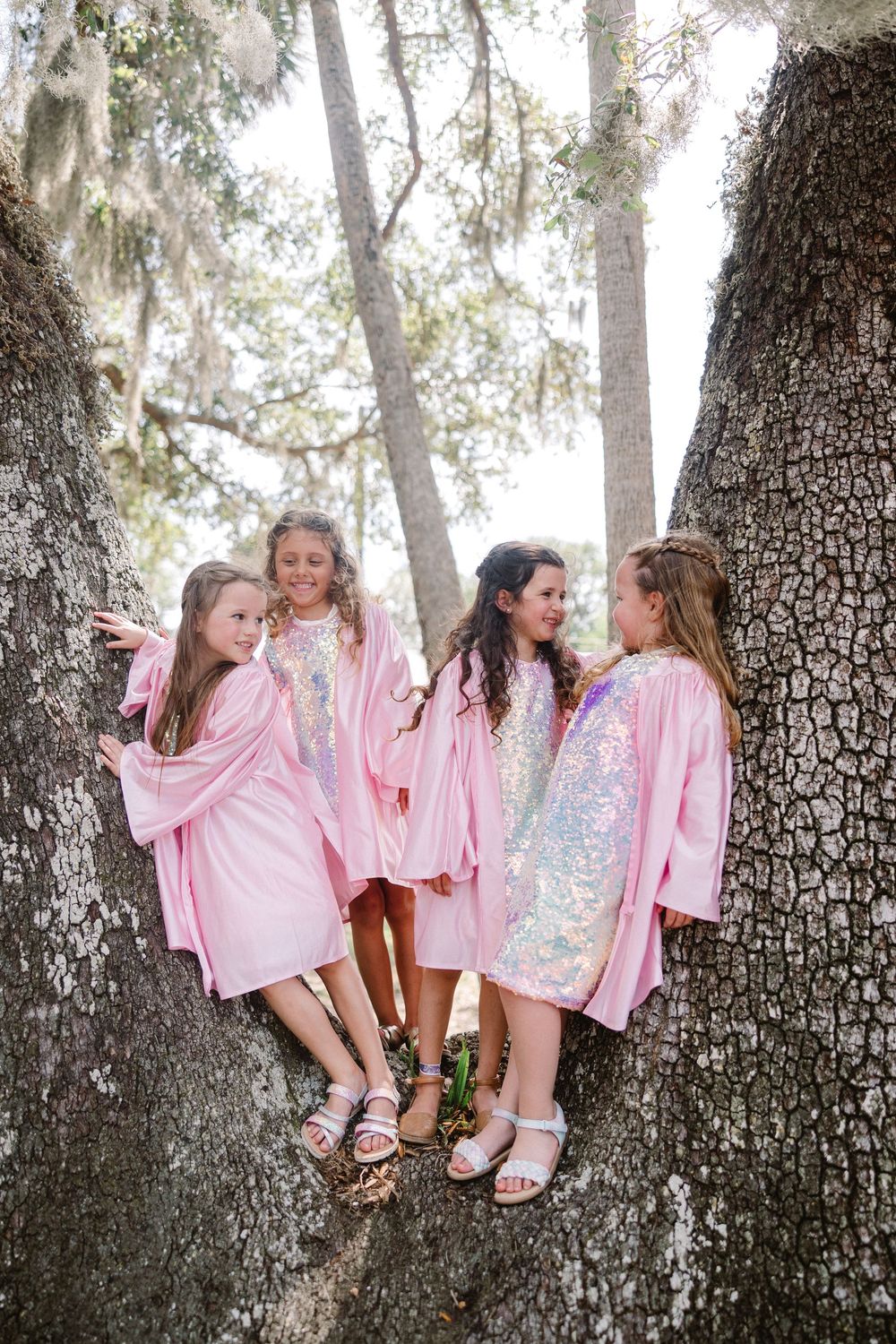 Four young friends in matching pink sparkly party dresses gather around a large oak tree trunk outdoors.