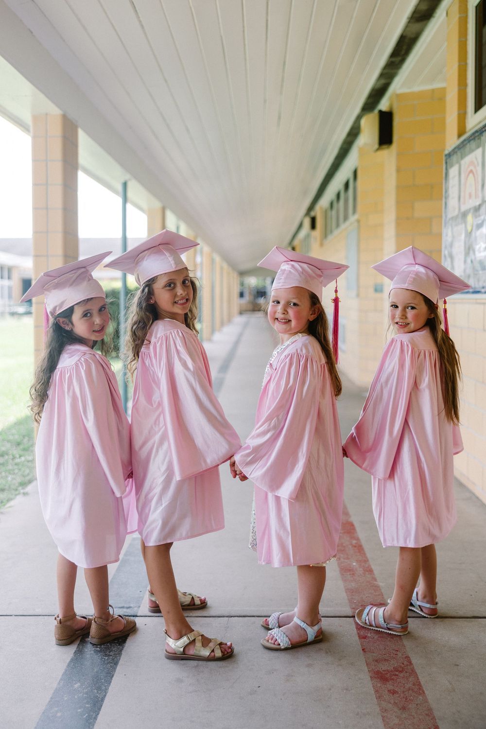 A sequence of shots showing young children in pink graduation caps and gowns playfully dancing in a school hallway.
