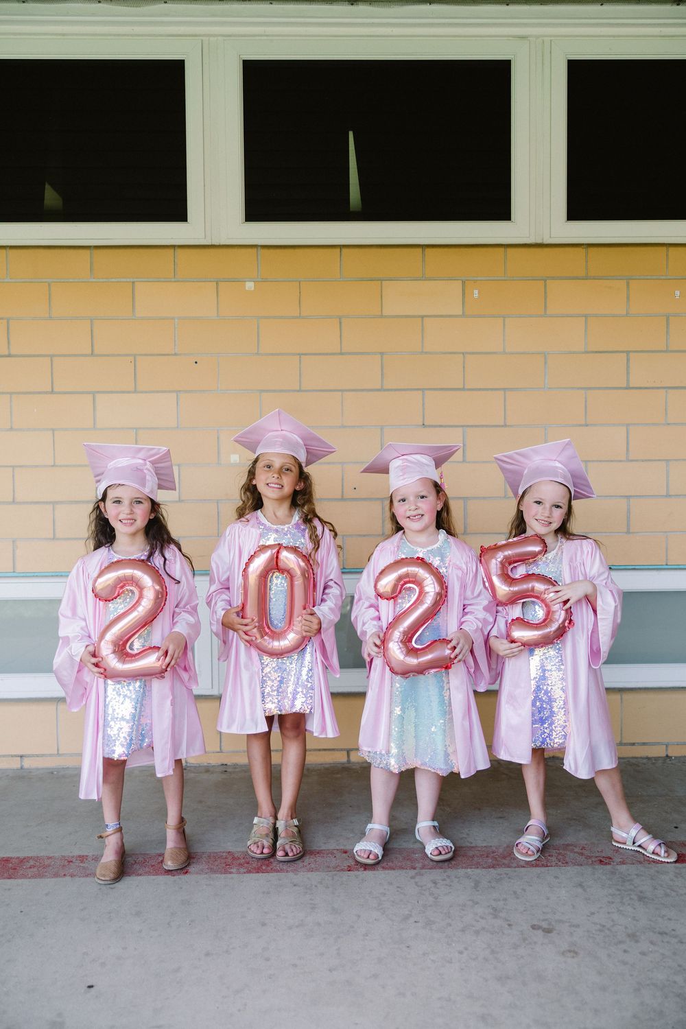 Four graduates in pink dresses sit together holding numbers 2023 while posing on concrete steps.