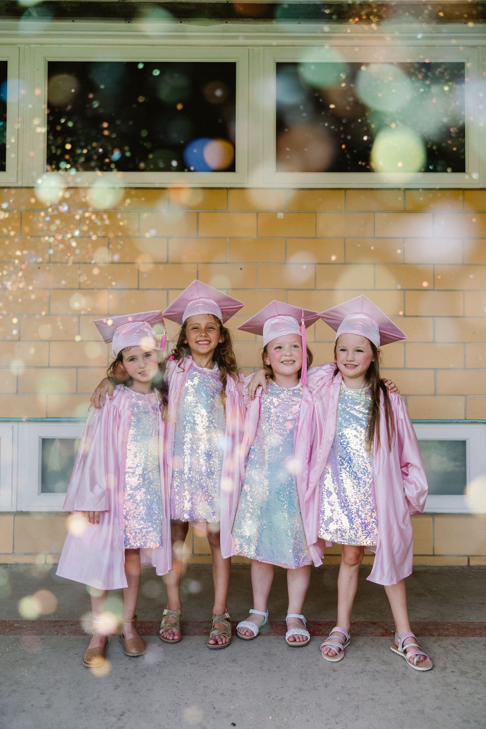 Four graduates wearing sparkly pink and silver dresses with matching pink caps stand together against a brick wall.