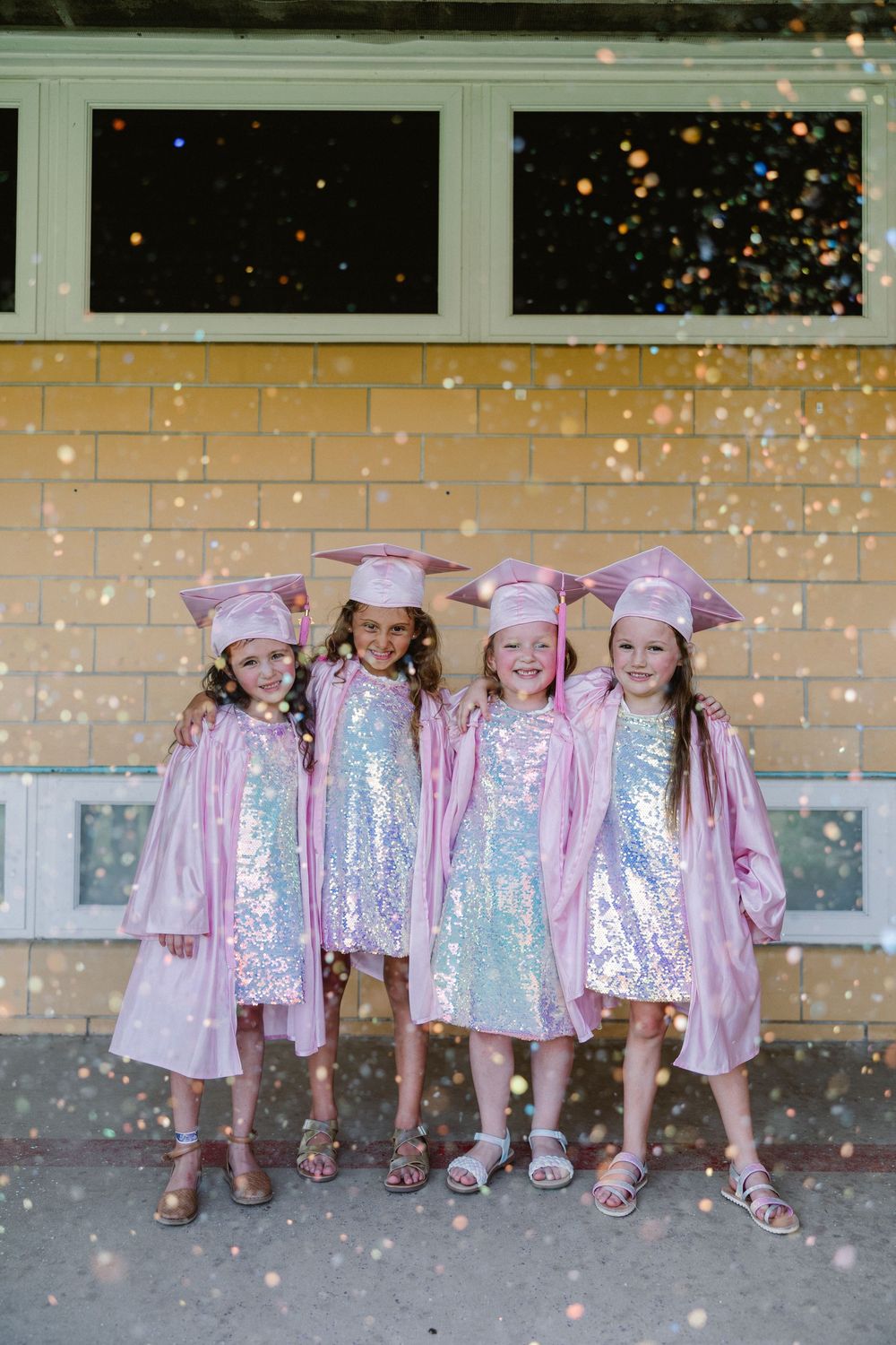 Group of small figures in matching shimmery pink outfits posing against a brick wall with sparkling confetti effects.