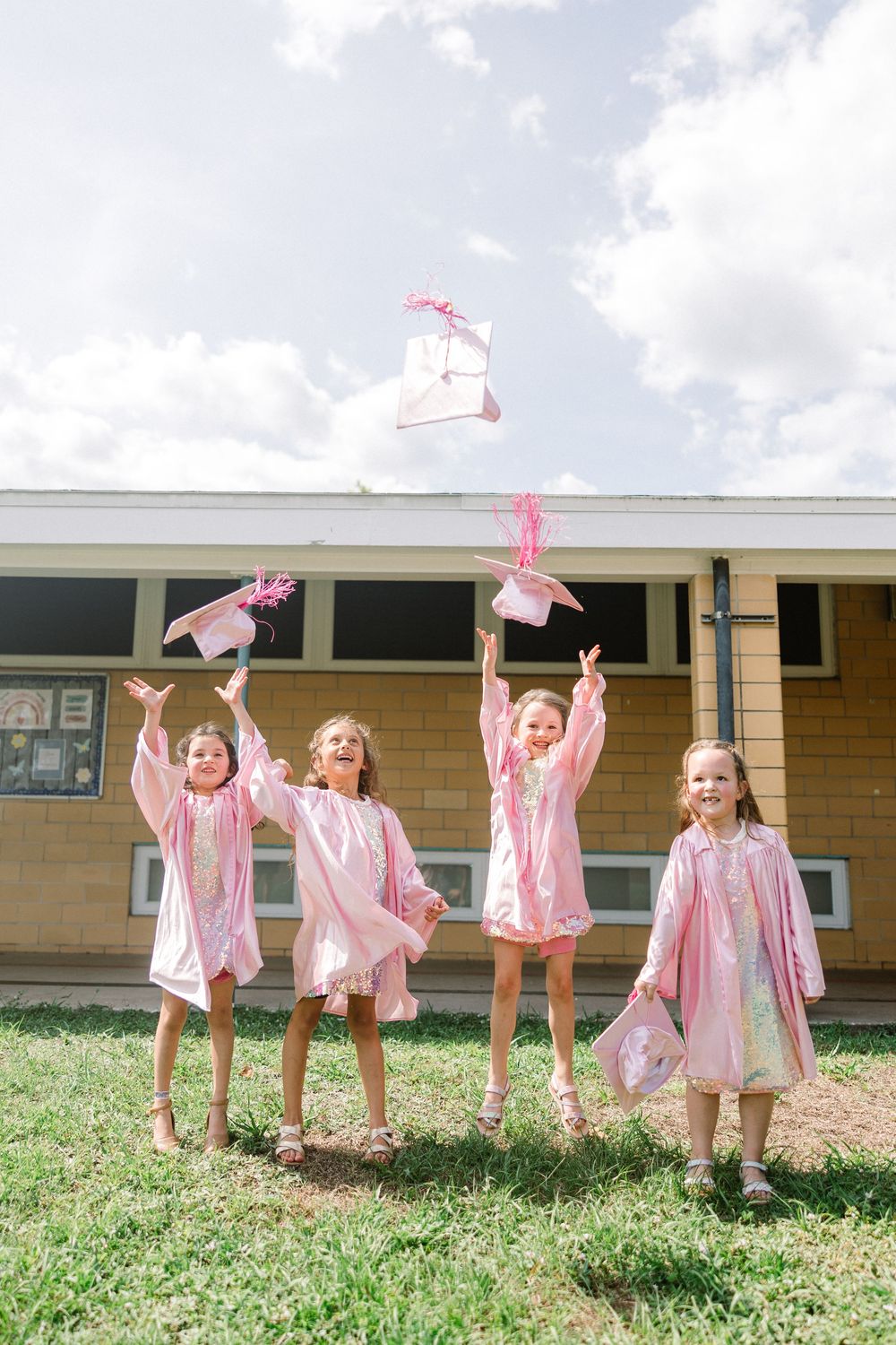 Four children in pink outfits joyfully throw items into the air while playing outside a building on grass.
