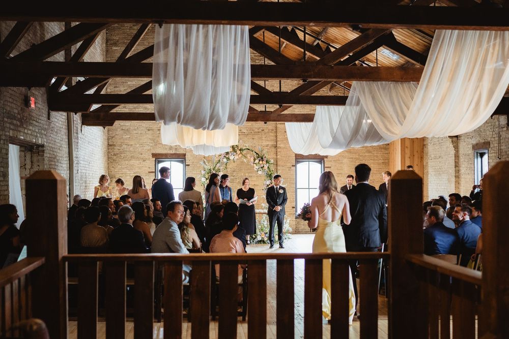 Elegant rustic wedding ceremony venue with white drapes hanging from exposed wooden beams and guests seated in rows.