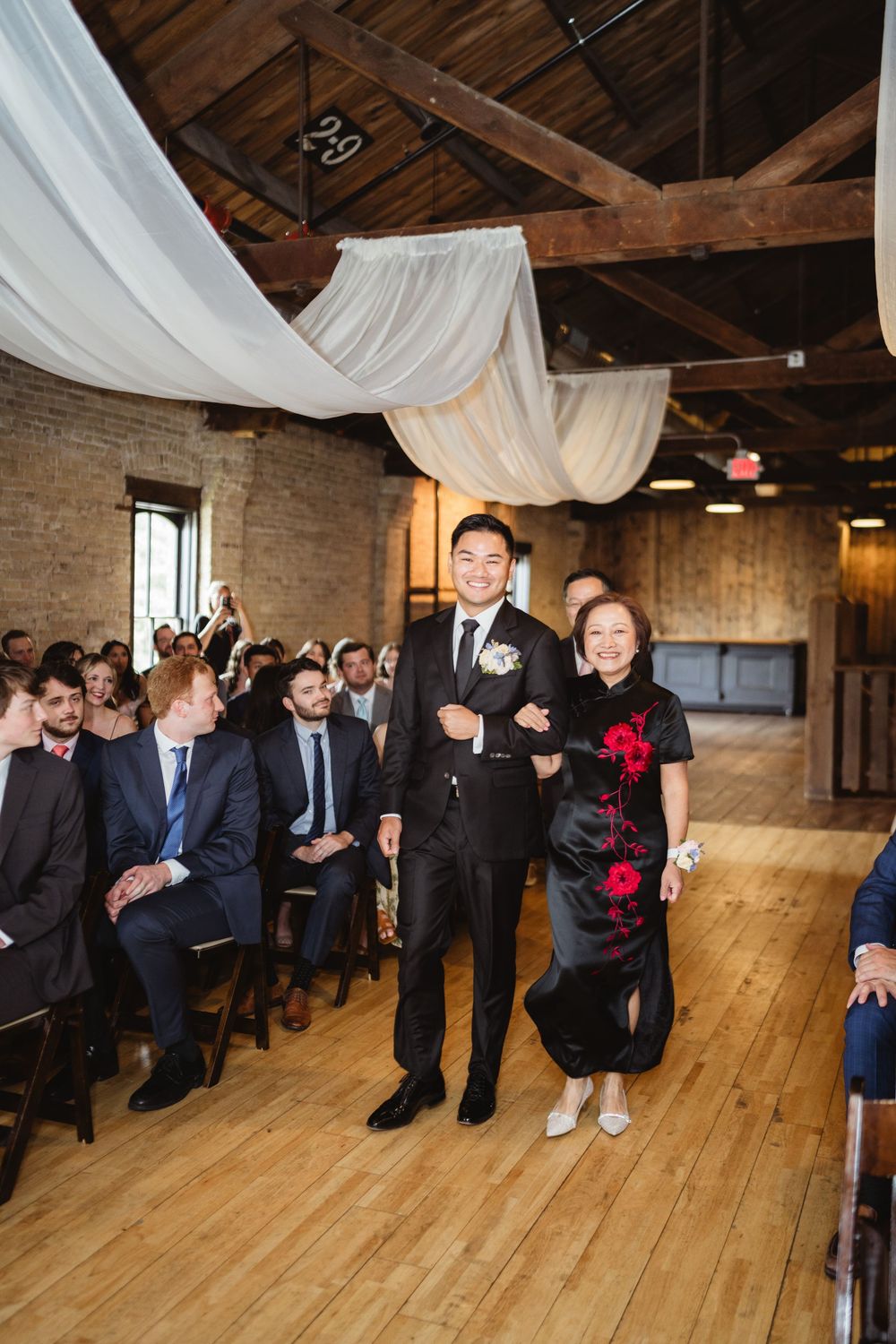 An indoor wedding ceremony in a rustic venue with exposed wooden beams, draped white fabric, and hardwood floors.
