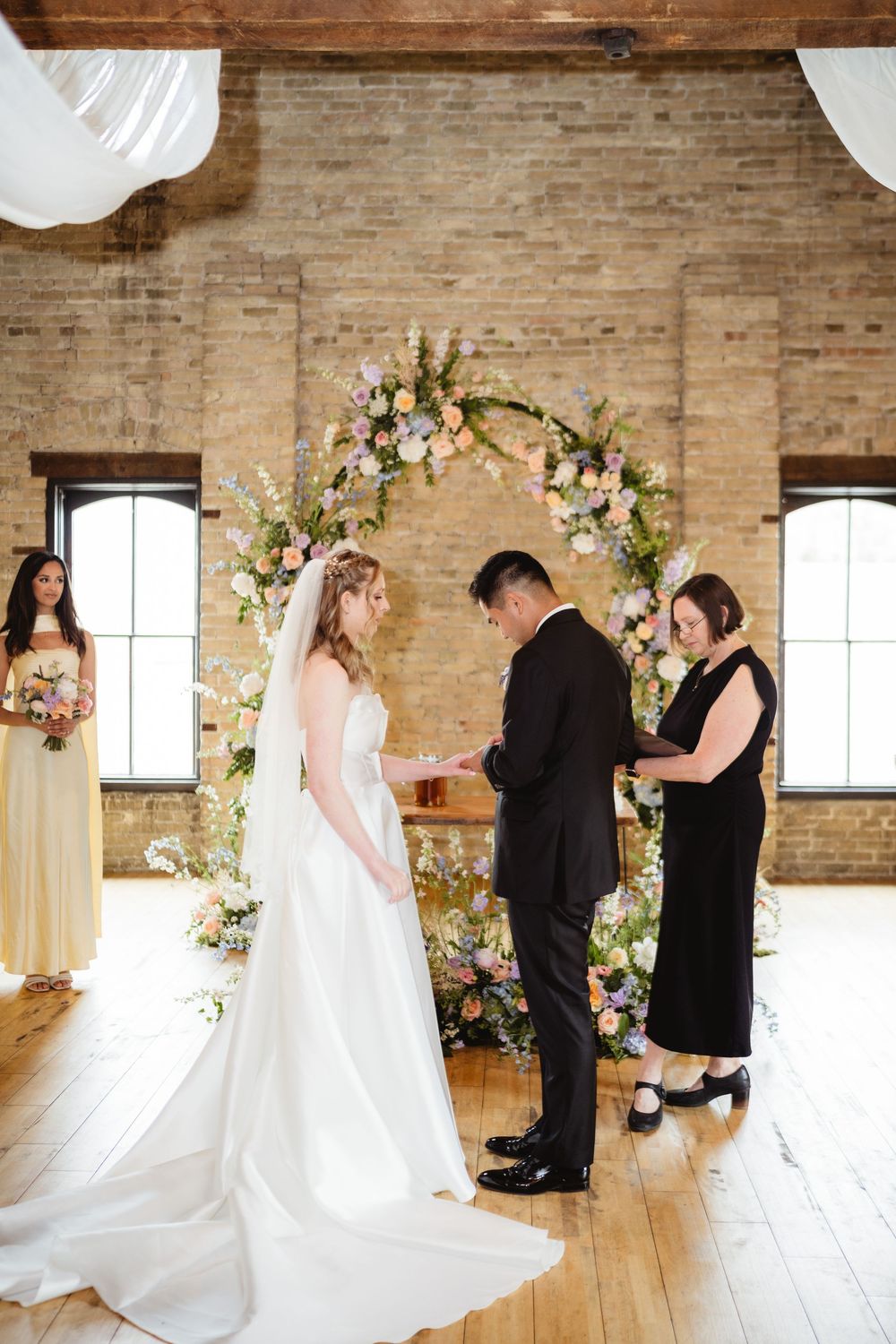 Wedding ceremony in rustic brick venue with wooden floors and floral arch during exchange of rings.
