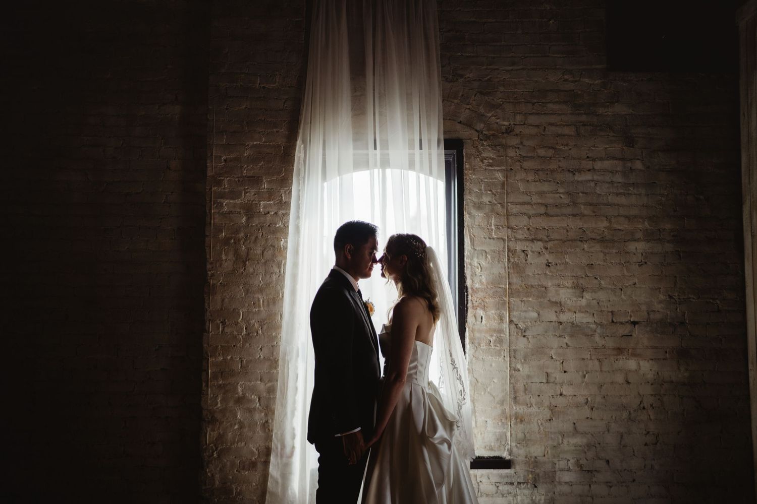 Silhouetted couple stands in romantic window light with brick walls creating a moody atmospheric scene.