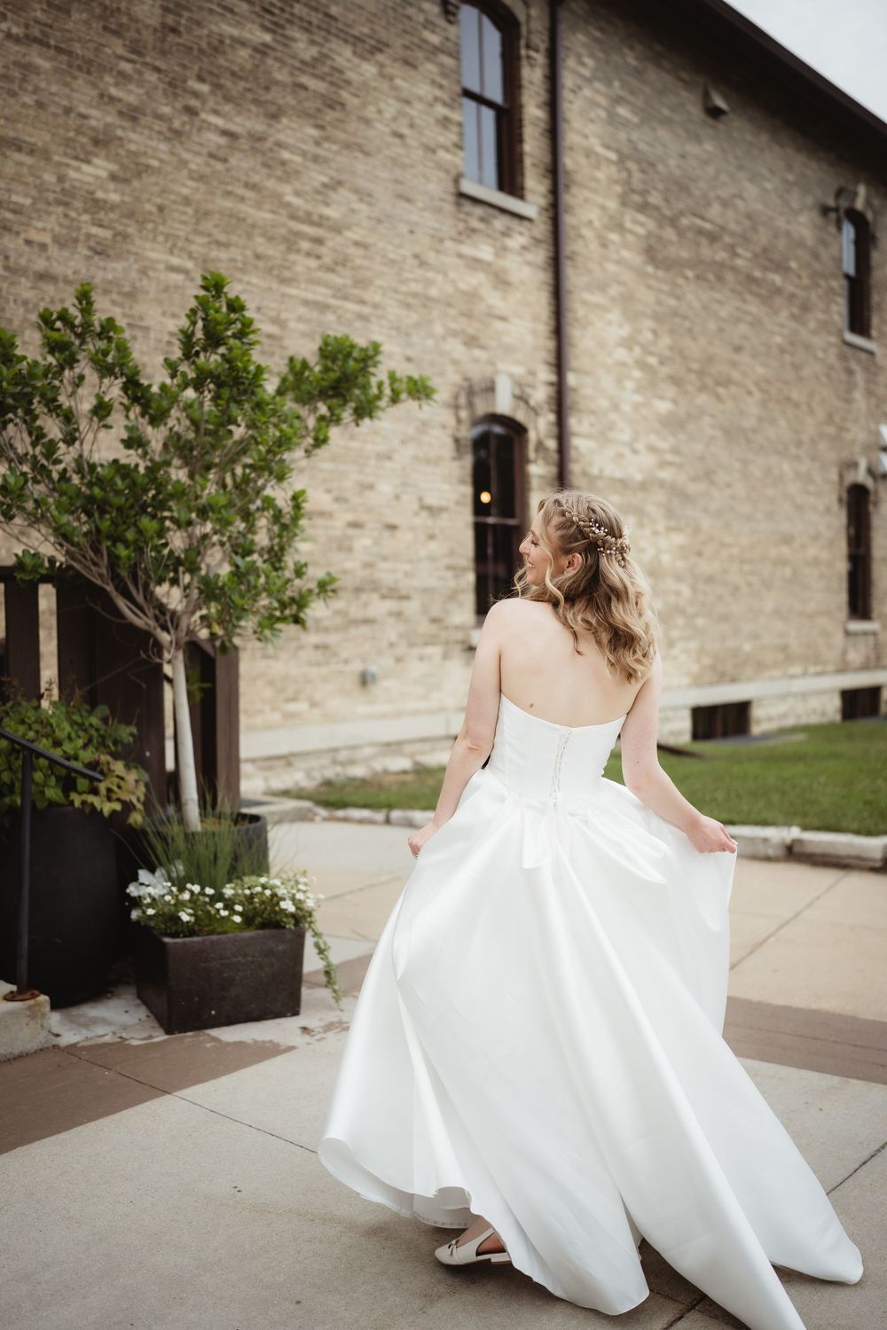 A bride in a flowing white wedding dress twirls gracefully outside a stone building on a sunny day.