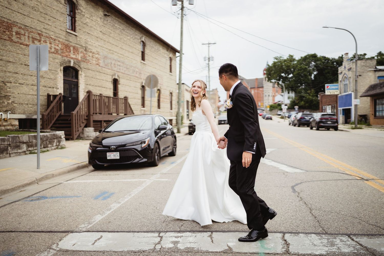Couple in wedding attire share a romantic moment on a city street in front of an industrial brick building.