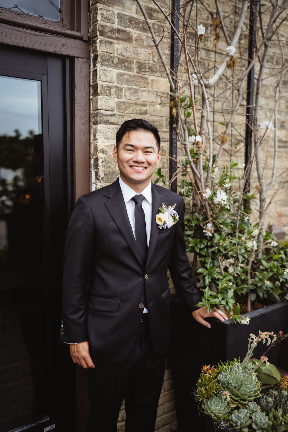 A person in a black suit and tie with a floral boutonniere stands smiling next to a brick wall with decorative plants.