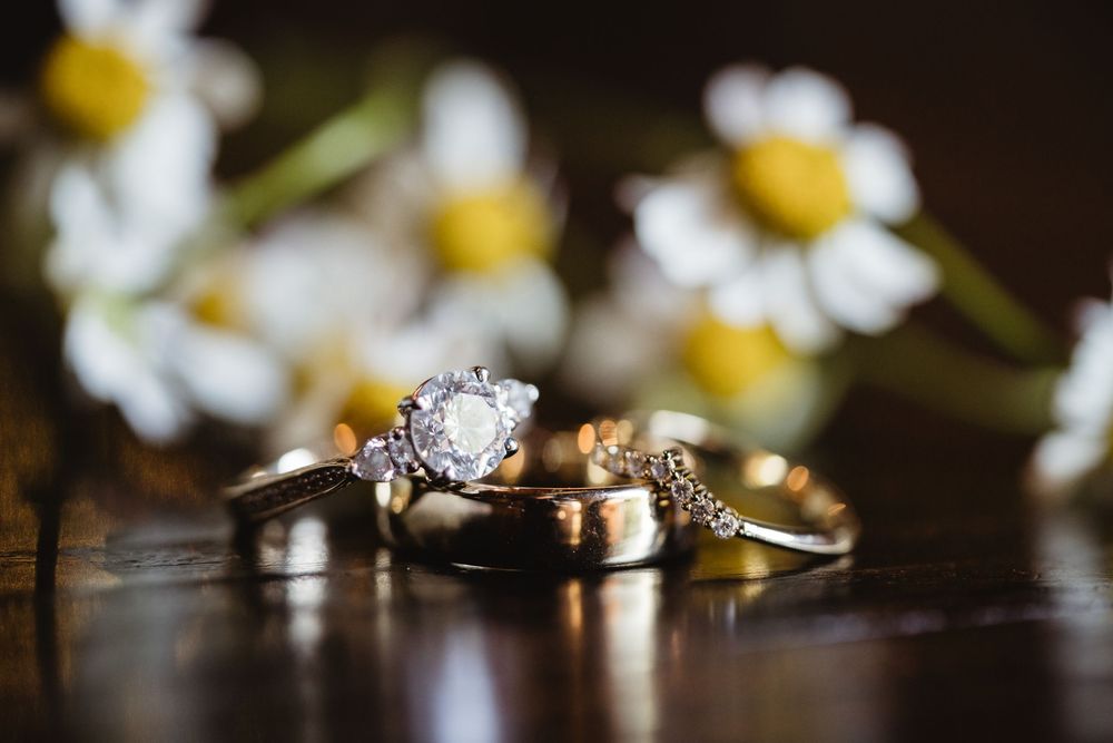 Wedding rings and engagement diamond ring artistically arranged with white daisies in background on dark reflective surface.