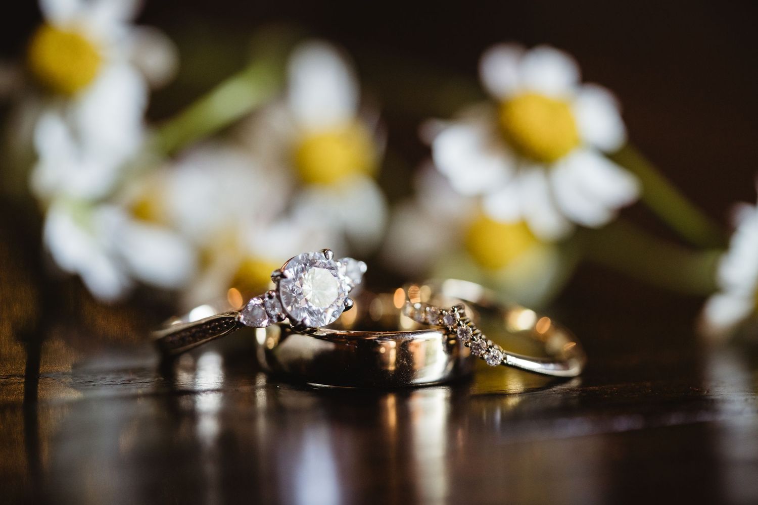 Wedding rings and engagement diamond ring artistically arranged with white daisies in background on dark reflective surface.