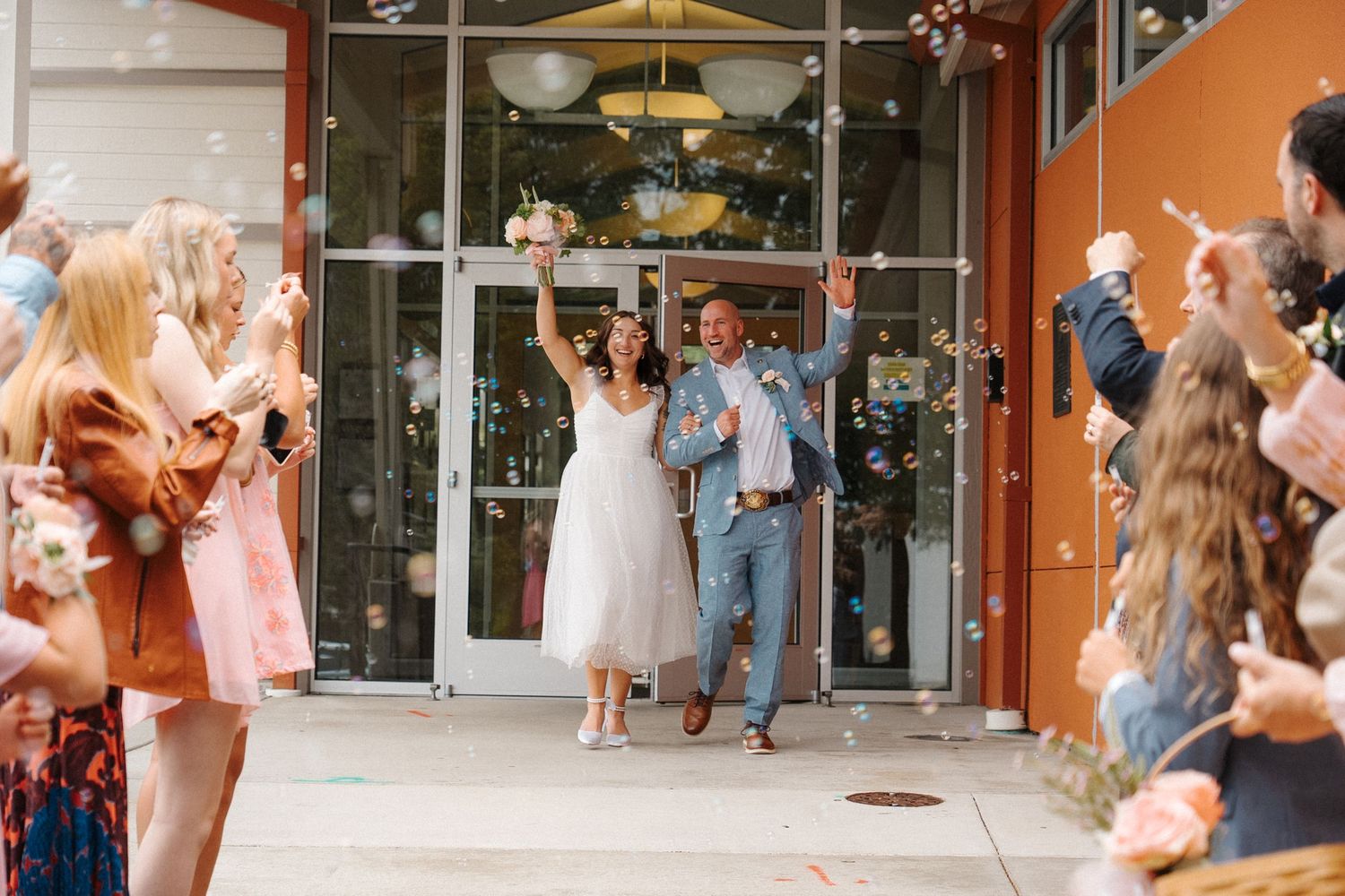 A happy couple exits a storefront venue while wedding guests celebrate and throw confetti on the sidewalk.