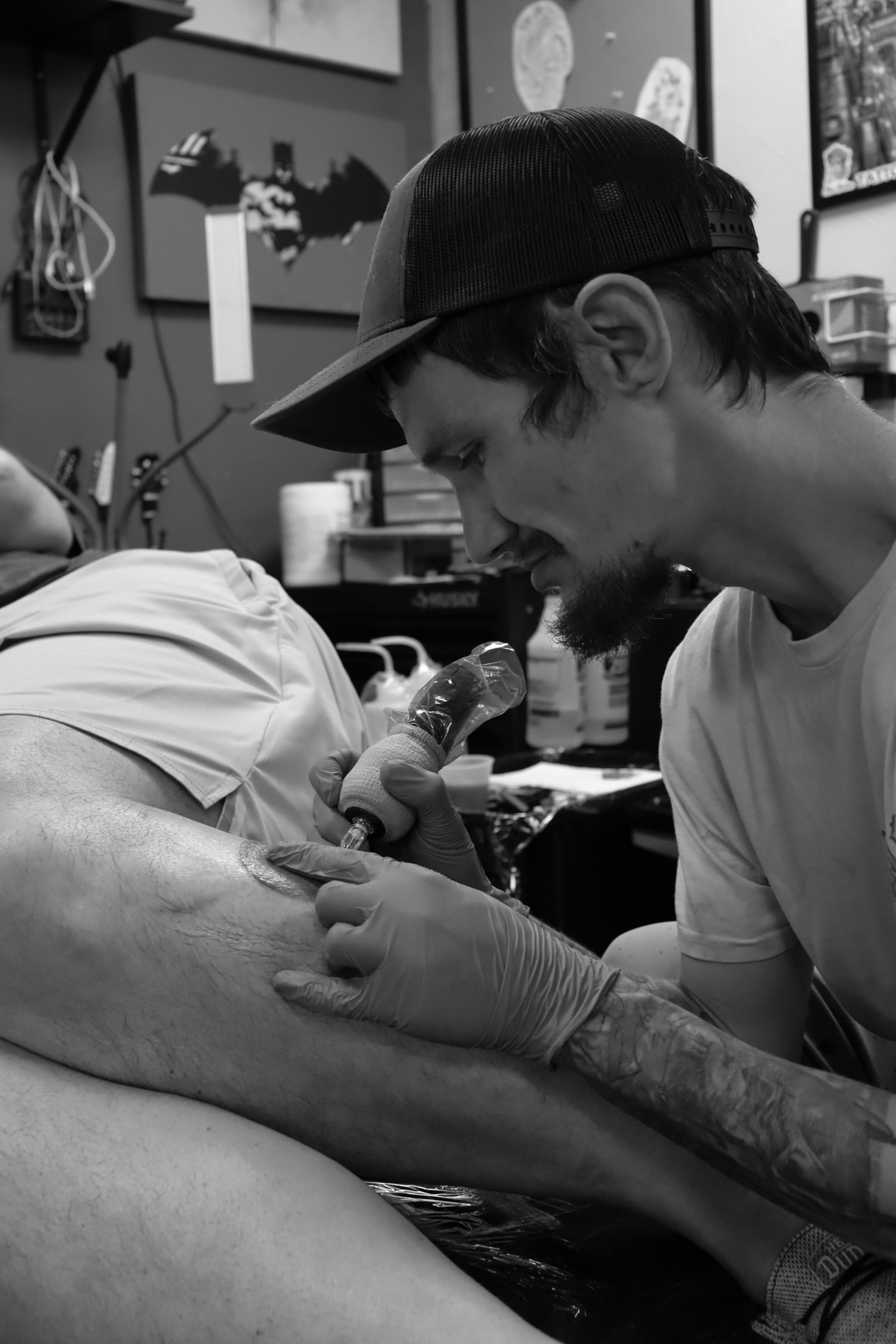 A tattoo artist in a black cap works on a client's arm in a black and white photograph taken at a tattoo studio.