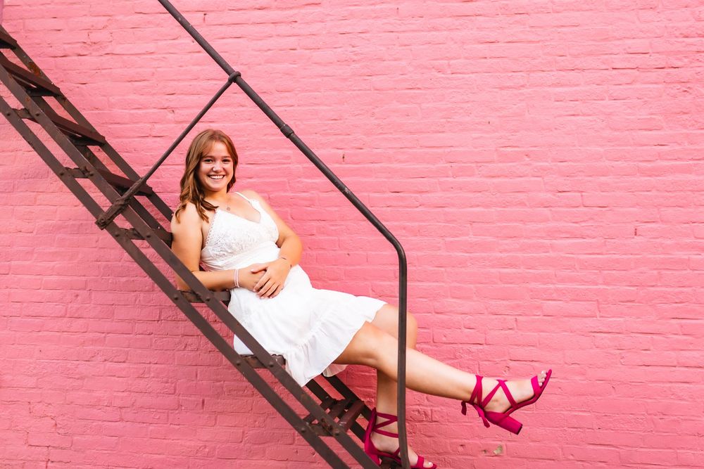 Portrait of a senior girl posing on the steps in front of a pink wall in Ionia Michigan.
