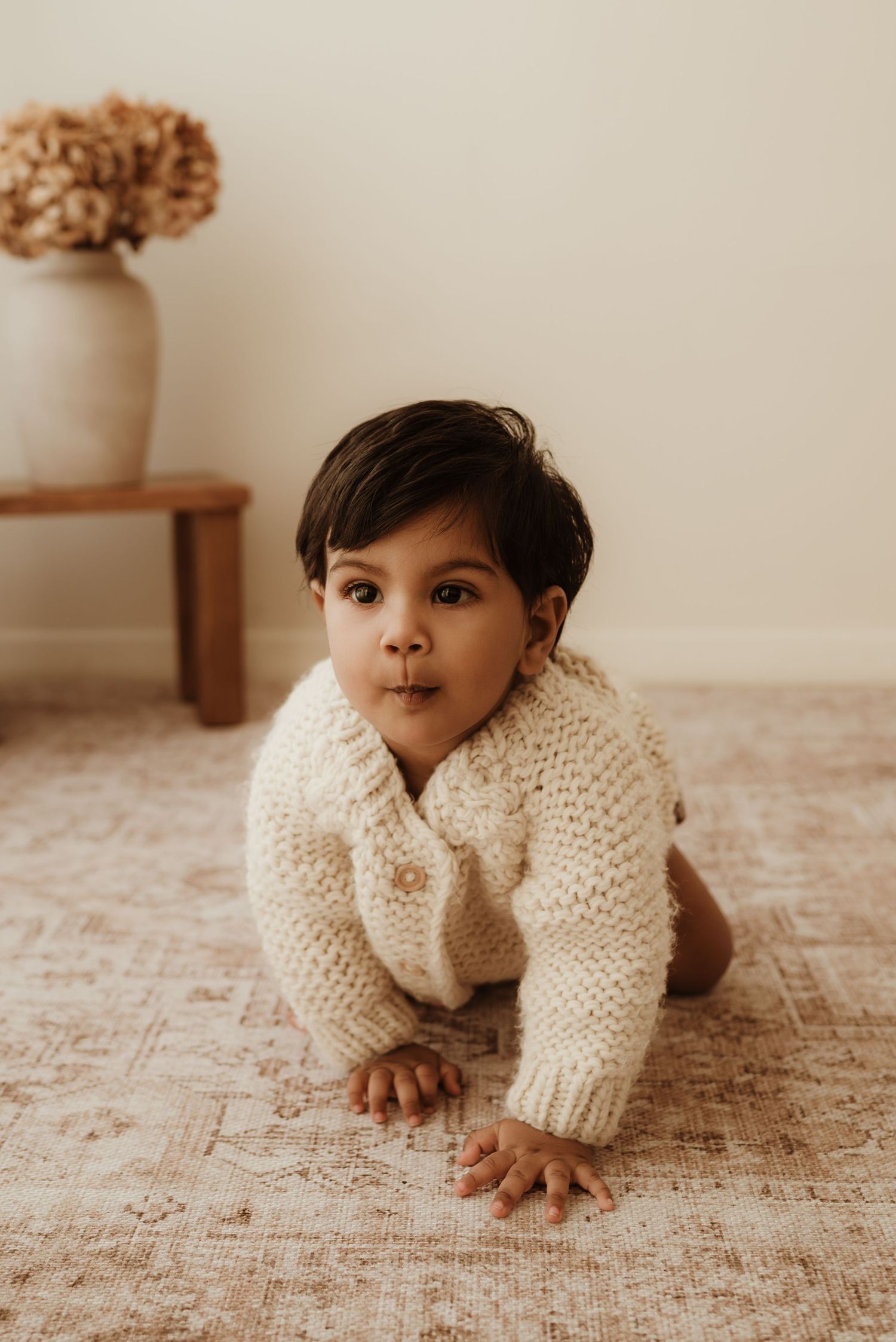 A baby wearing a white ruffled dress sits on a textured cream blanket in warm natural lighting.