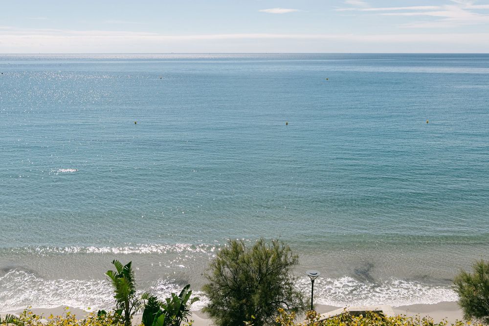 Panoramic view of a turquoise ocean coastline with gentle waves rolling onto a sandy beach under a bright blue sky.