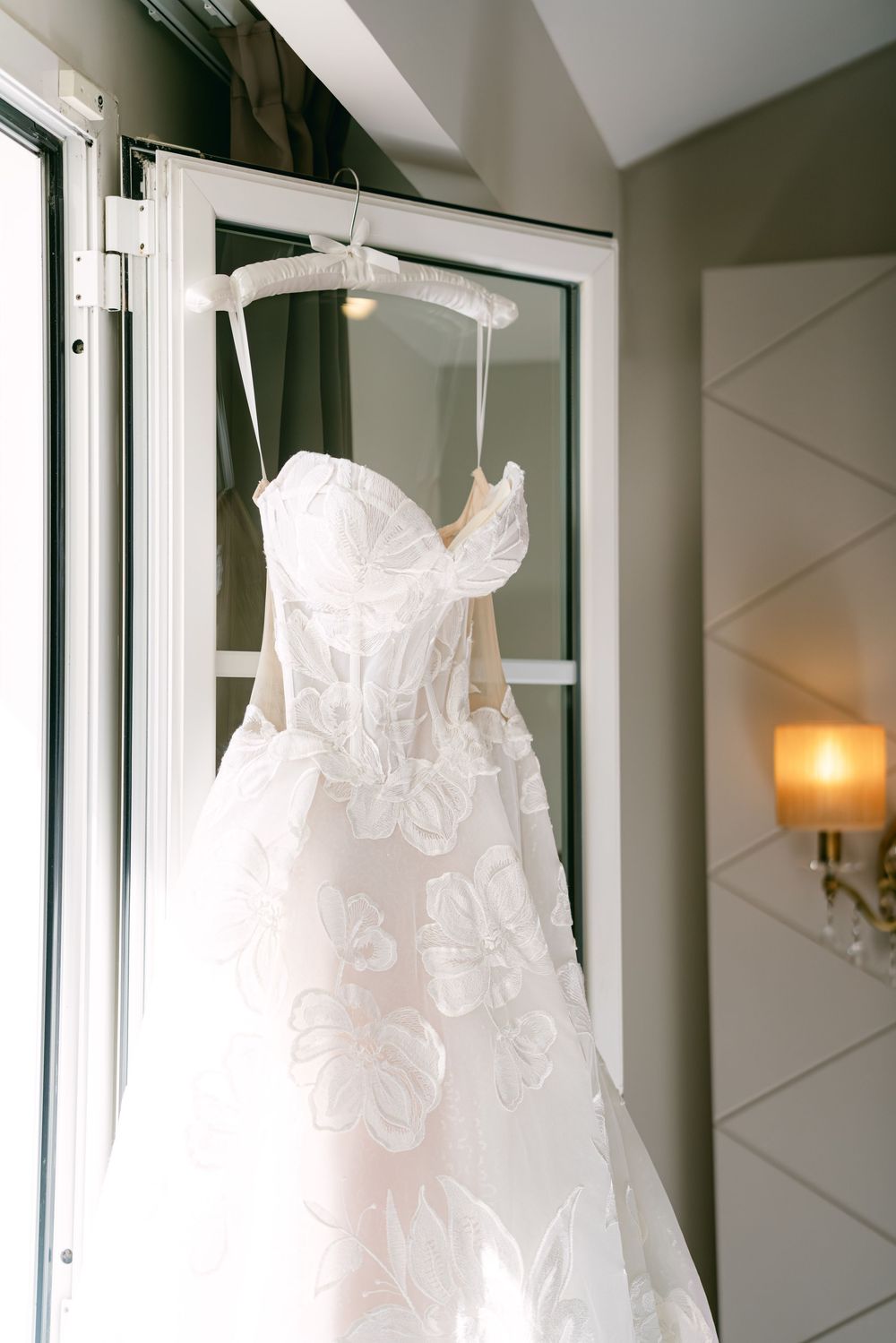 White wedding dress with delicate lace details hangs elegantly against a mirrored wall in a bridal suite.