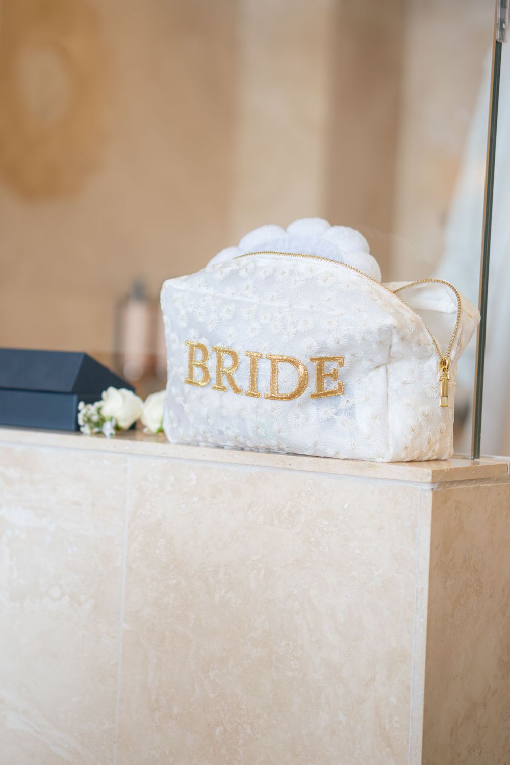 A white lace cosmetic bag with gold 'BRIDE' text sits on a marble surface next to baby's breath flowers.