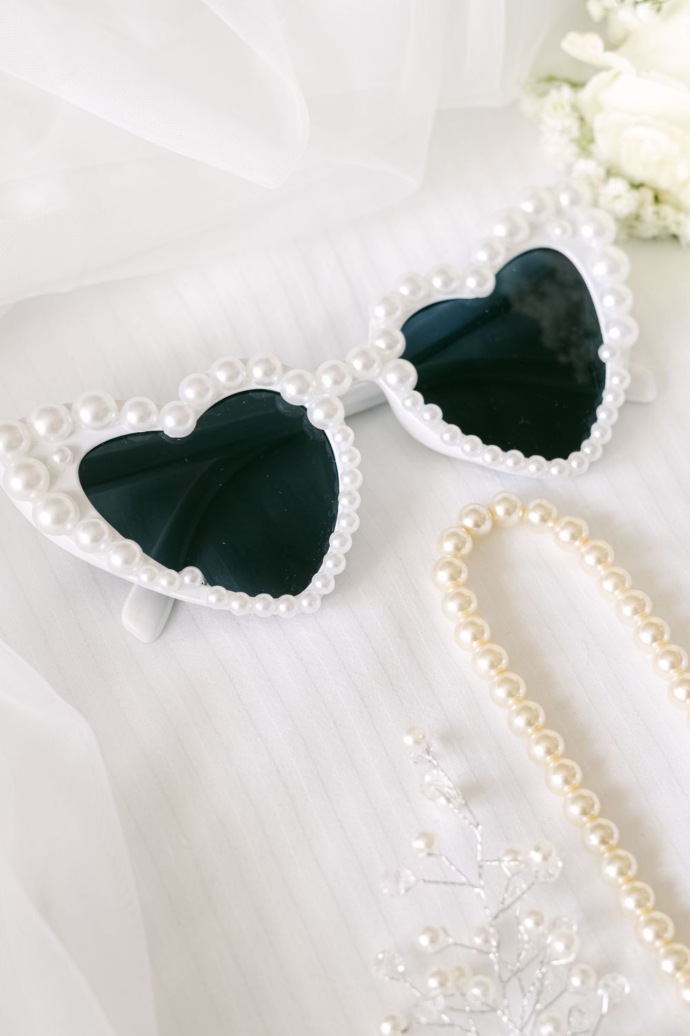 Heart-shaped sunglasses with pearl trim lay elegantly beside a delicate pearl necklace on white fabric.