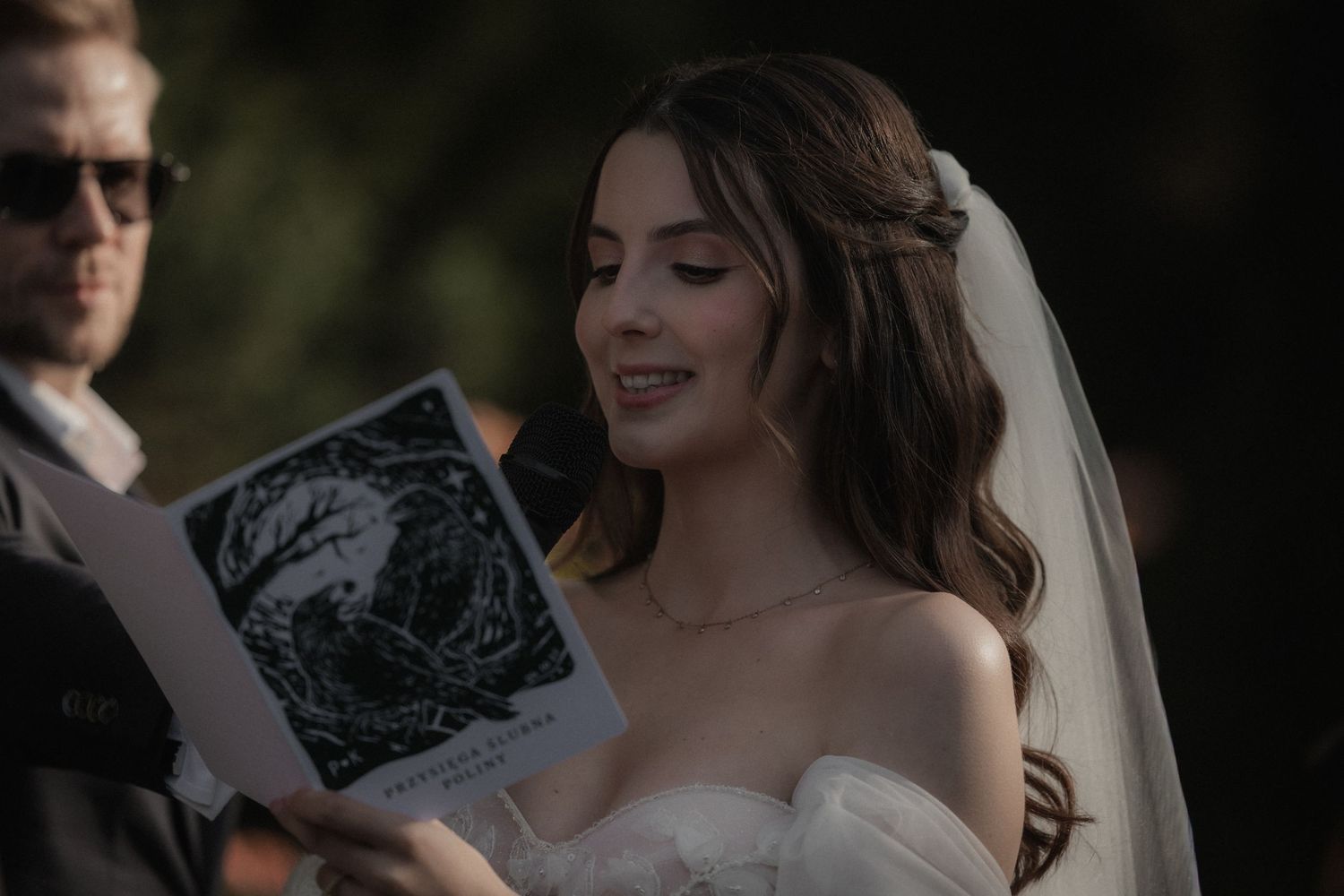A bride in a white strapless wedding dress smiles while looking at a document during an outdoor ceremony.