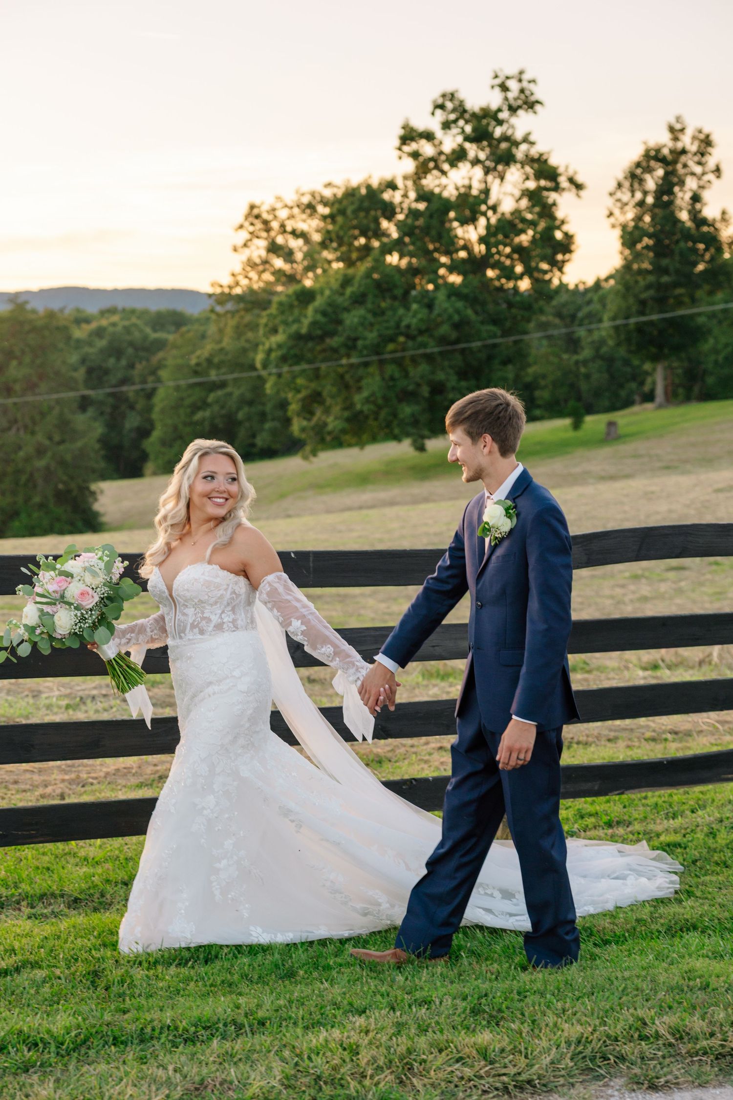 A couple in wedding attire playfully runs and spins on a grassy hill with a wooden fence at sunset.
