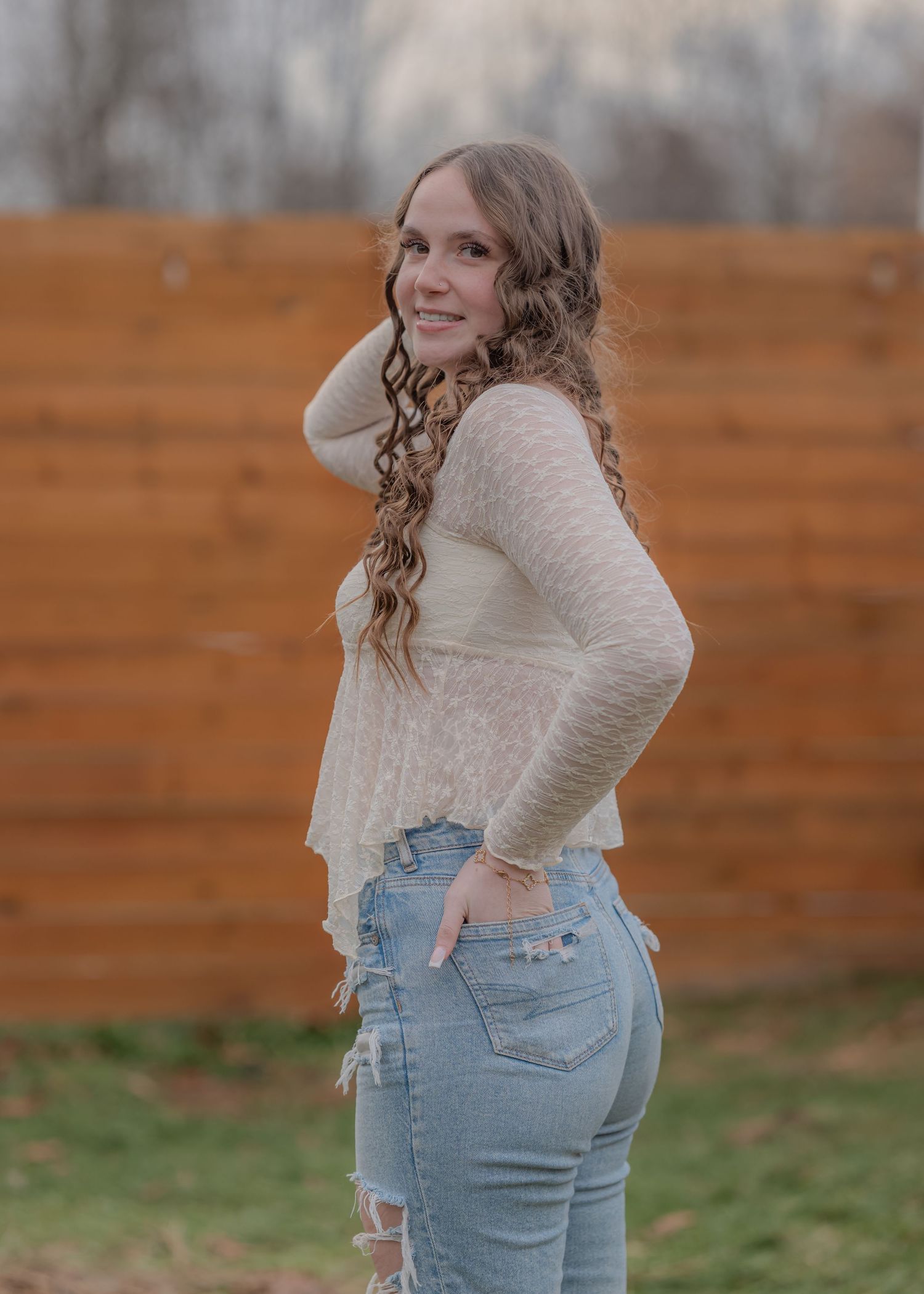 Side view of a person wearing a cream sweater and light blue jeans posing against a wooden fence background.