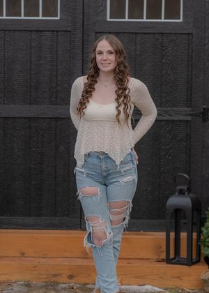 A person wearing a cream lace top and distressed blue jeans poses against a dark wall on a wooden deck.
