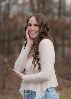 A person in a white lace top and jeans poses casually in a wooded outdoor setting during autumn.