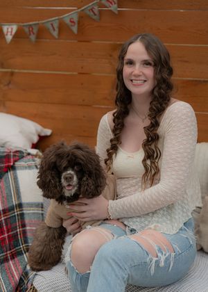 Person in cream sweater and distressed jeans cuddling with small brown dog on plaid blanket.
