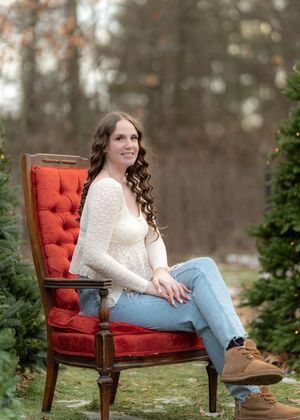 Person relaxing in vintage red velvet chair surrounded by evergreen trees in outdoor setting.