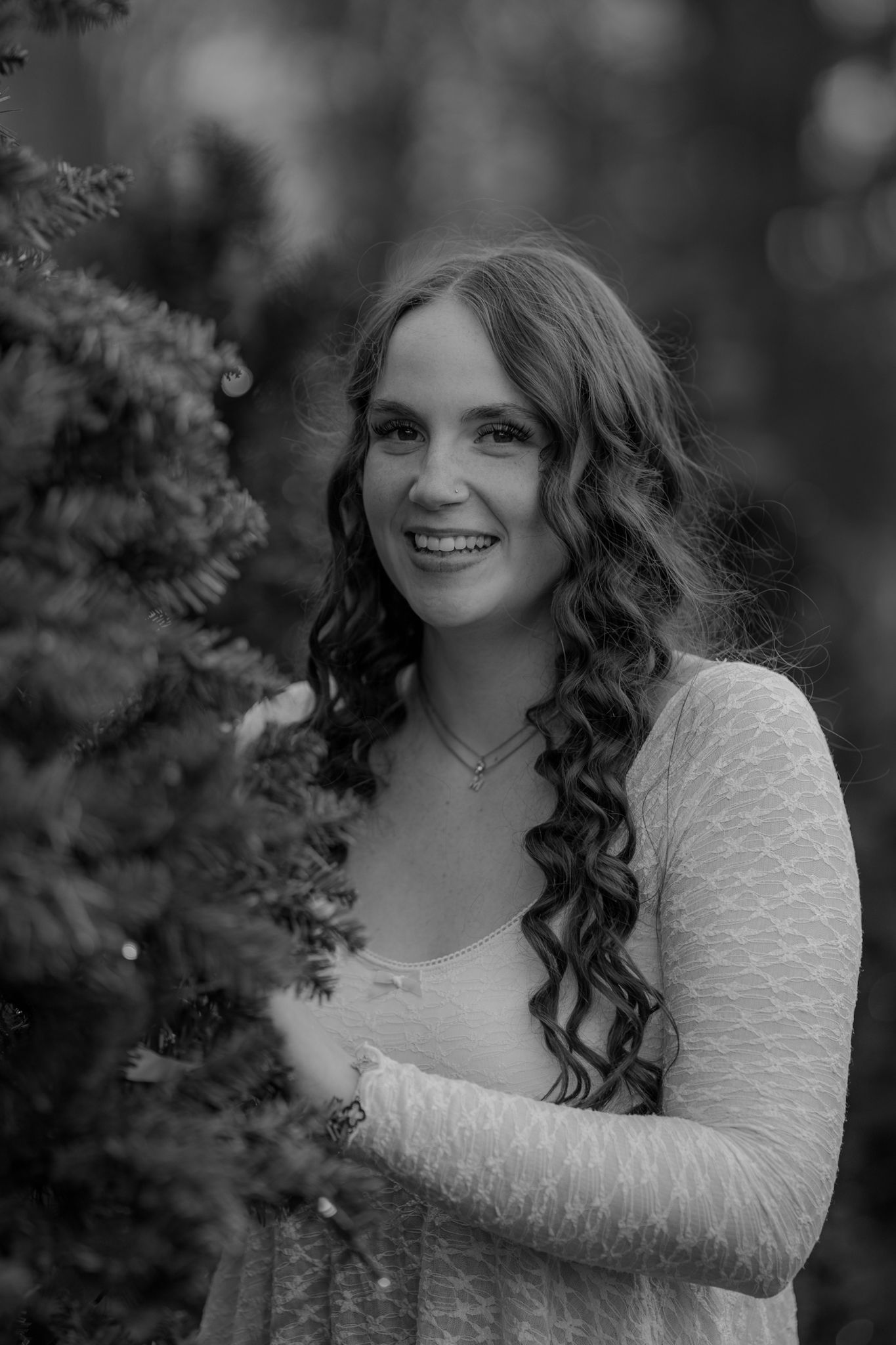 Black and white portrait of a person with long curly hair and a lace dress smiling outdoors near foliage.