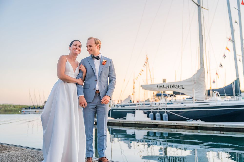 A couple celebrates their wedding day on a scenic dock with boats and water in the background at sunset.