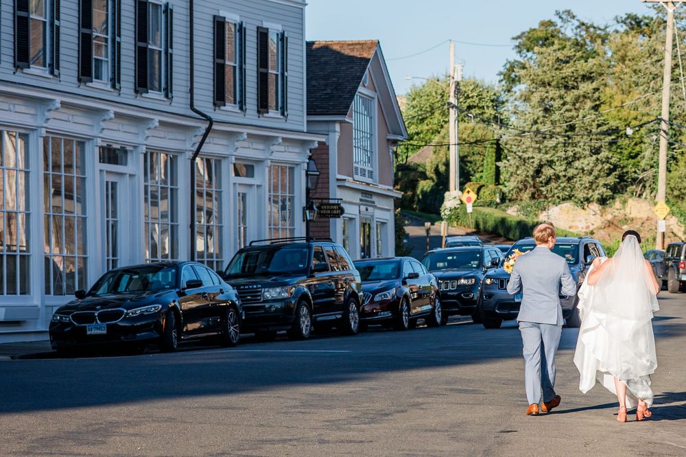 Two people walk down a street lined with parked cars and historic buildings in a quaint New England town.
