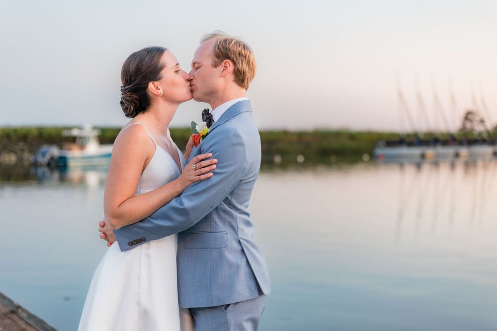 A couple in wedding attire shares a romantic kiss by a waterfront dock at sunset.