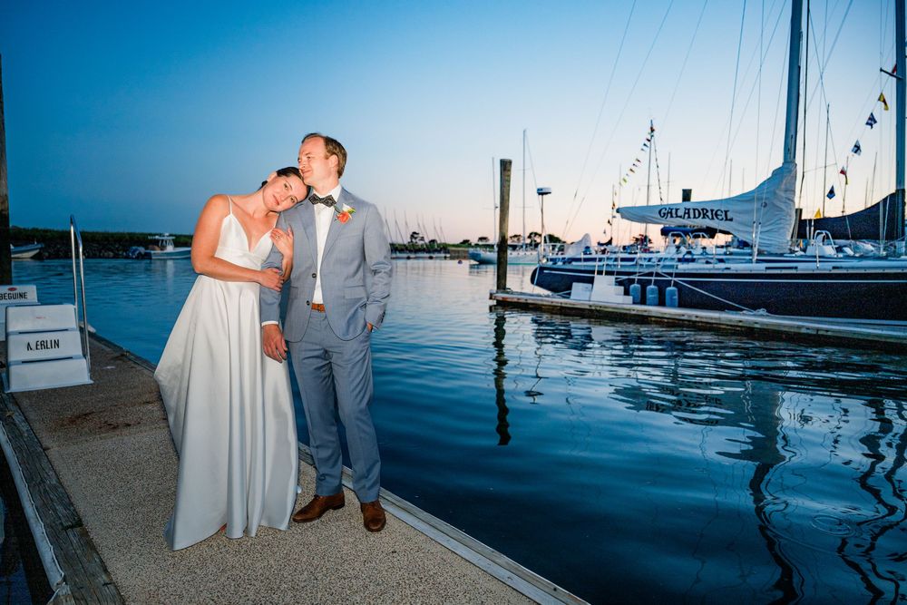 A wedding couple stands together at sunset on a dock with boats and sailboats in the harbor background.